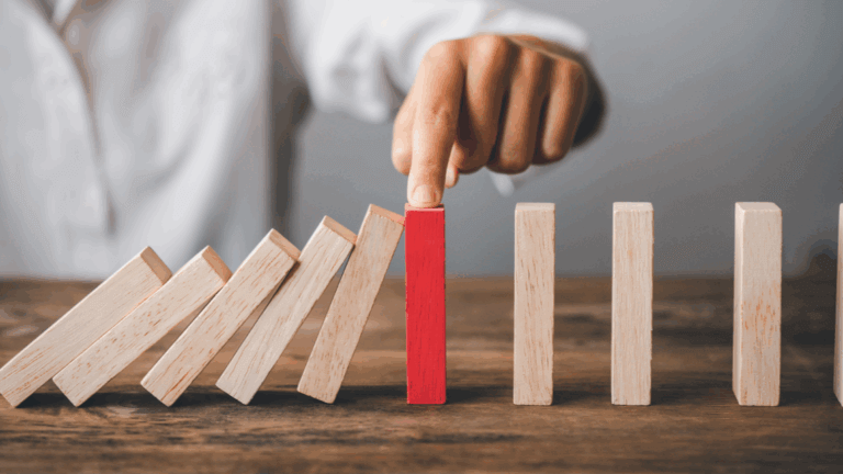 Hand stopping red wooden block from falling in domino chain, preventing cascade effect.
