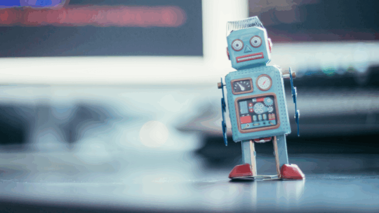 A small robot standing on a desk in front of computer monitors displaying website code.